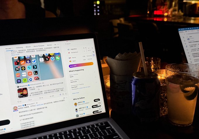 A laptop displaying a Twitter/X feed sits on a bar counter next to drinks, suggesting a relaxed work session—fitting the post's playful tone of "drinking on the job" while promoting Bitget's full suite of services for collaboration.
