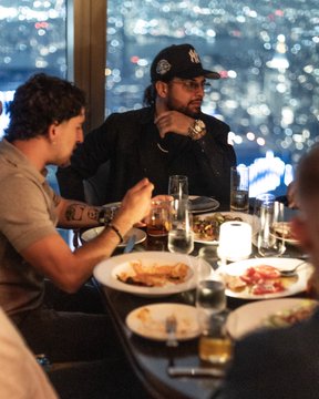 Two men sit at a dinner table with city lights visible through the window behind them, with one wearing a black cap and glasses. The caption "this is me / this is my doginal dog" humorously frames the scene as the man treating his dining companion like a beloved pet.