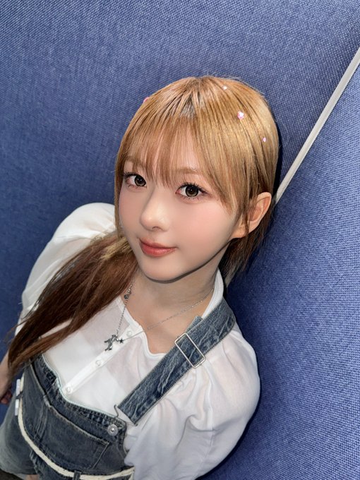 A young woman with light brown hair and bangs poses against a blue fabric background, wearing a white blouse under denim overalls.