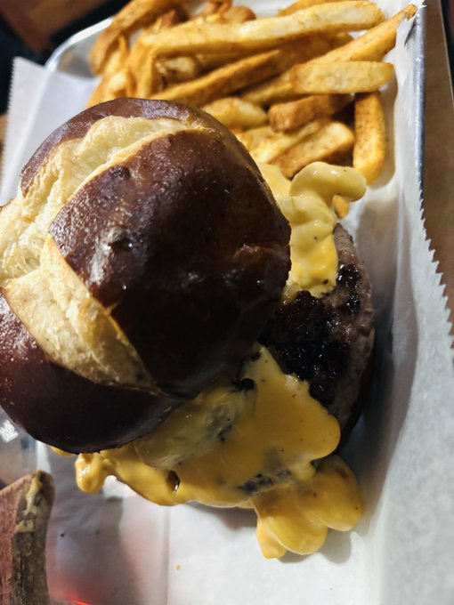 A close-up of a cheeseburger with melted yellow cheese on a pretzel bun, served with seasoned french fries on white paper.