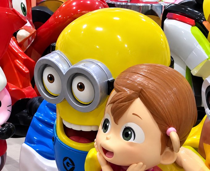 A close-up photograph of glossy amusement park ride vehicles featuring 3D figures of a Minion and a brown-haired cartoon girl, with other colorful characters visible in the background.
