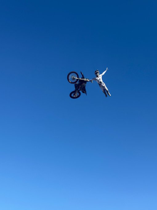 A motocross rider catches big air high against a clear blue sky, performing a no-hander stunt with arms outstretched while the bike tumbles alongside, perfectly matching the "FULL SEND" energy referenced with @johnknopf.