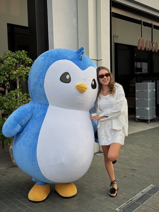 A woman in a white shirt, white skirt, and sunglasses poses next to a large blue and white penguin mascot costume outside a building.