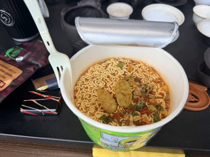 A cup of instant noodles with chili seasoning and green vegetables, accompanied by a plastic fork on a dark surface near an airplane seat.