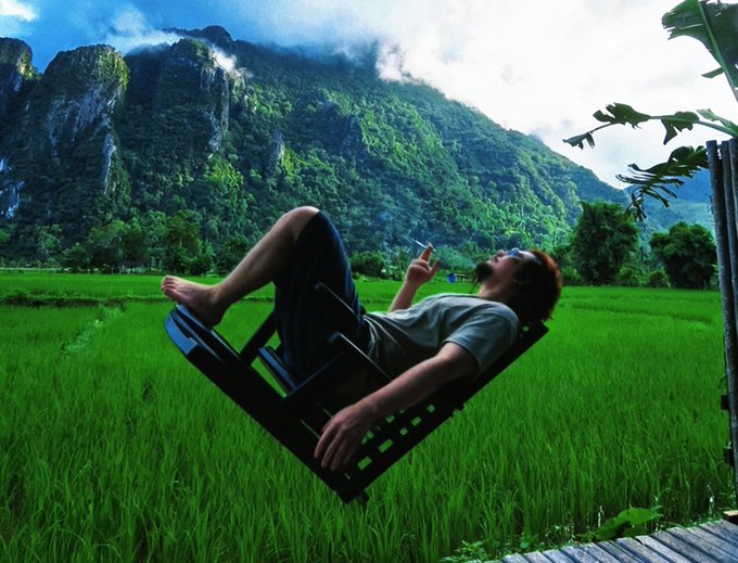 A person relaxes on a black lounge chair balanced in a vibrant green rice field with dramatic mountains in the background.