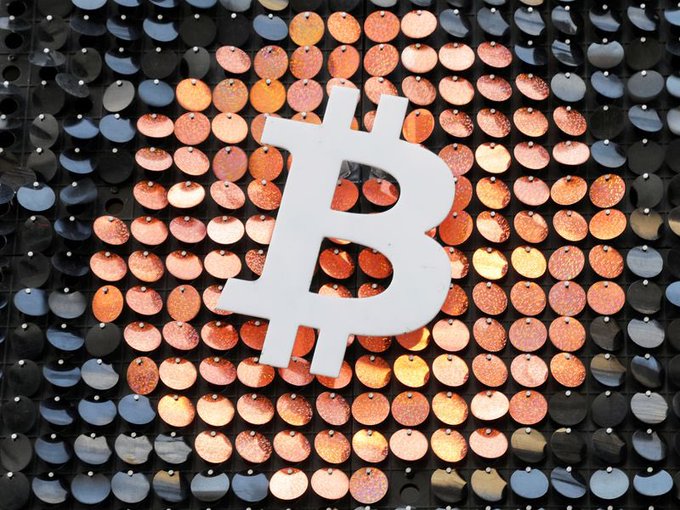 A large white Bitcoin (₿) logo mounted in front of a backdrop made of reflective copper and dark metallic circular discs.