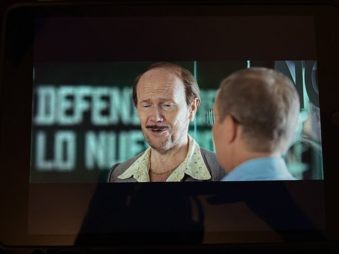A man watches a scene from the Spanish film "Torrente" on his iPad, with the partial text "DEFEN... LO NU..." visible on screen behind a mustachioed character, capturing his relaxed multitasking night of streaming a movie while geomining with Coin app.
