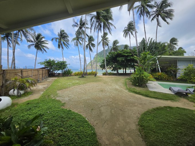 A tropical backyard view featuring palm trees, a grassy lawn with dirt paths, a bamboo fence, and the ocean visible beneath a cloudy sky with mountains in the distance.
