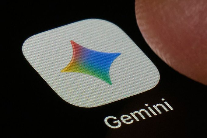 A close-up macro photograph of a smartphone screen displaying the colorful Gemini app icon and its name, with a finger visible in the background.