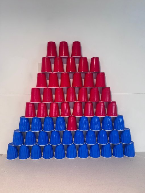 Red and blue plastic cups are stacked in a tall pyramid formation against a plain wall.