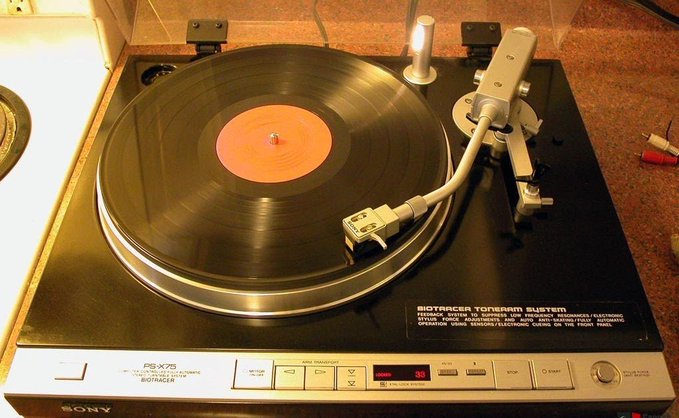 The image features a black turntable with an orange record, placed on top of a countertop or table. There are two red speakers positioned next to the turntable, which appear to be connected to it. The background seems to be a kitchen area, as there is a stove visible in the scene. A caption below the image reads: "Jokes on you my system is entirely analog." This phrase is likely a reference to the analog nature of the turntable and speakers, suggesting that they are not digital or electronic devices but rather traditional music equipment.