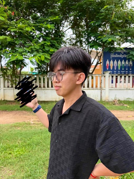 A young man with glasses in a black shirt poses outdoors, with a censored black scribble covering the object in his raised hand. Behind him, a "facebook" sign lists various country icons, playfully illustrating his post about avoiding the leaderboard rankings.