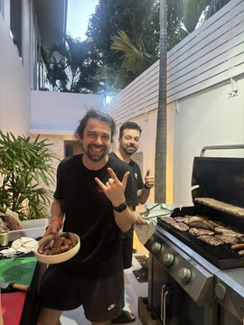 Two bearded men, including @shimas_sol, stand by a smoking grill during a Brazilian BBQ night at Island DAO's @RedotsClub event, proudly showing off fresh-cooked meat and giving the rock-on hand sign.