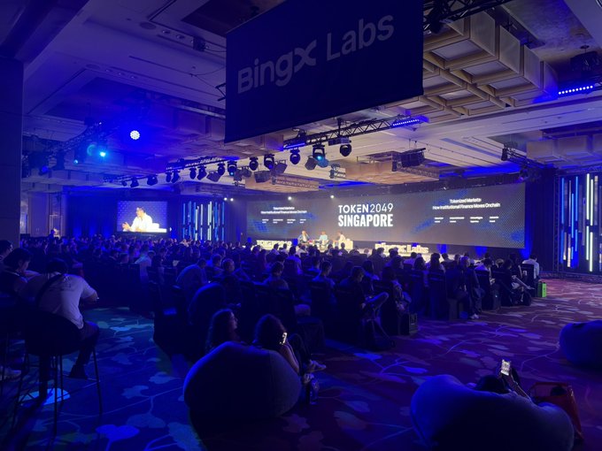 Attendees gather at the BingX Labs-sponsored Token 2049 Singapore conference, where the main stage is set for upcoming panels, including one titled "The Past, Present and Future of Robinhood Chain." The scene captures the anticipation just before the speaker takes the BingX stage to discuss the evolution of Robinhood Chain.