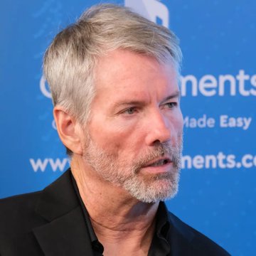 A close-up portrait of a gray-haired, bearded man in a dark shirt speaking at what appears to be a press event with blue branding visible in the background.