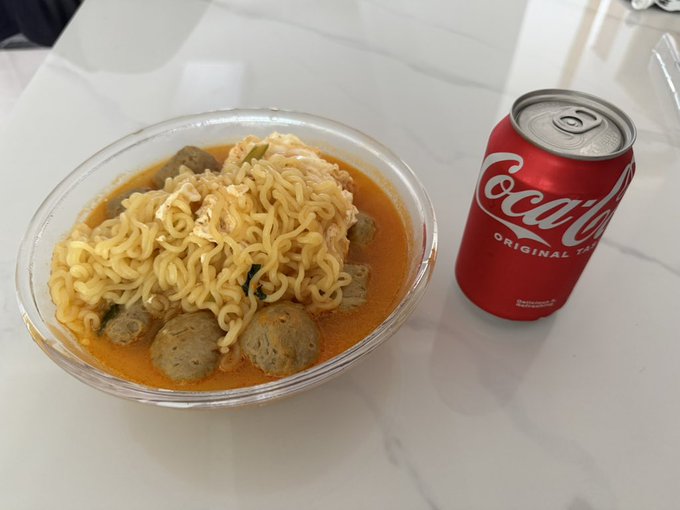 A plastic bowl of instant ramen noodles with meatballs in orange broth sitting on a marble counter next to a can of Coca-Cola Original Taste.