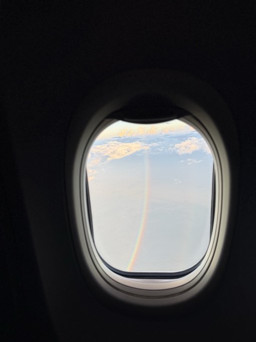 A view through an airplane window showing a vibrant rainbow stretching down from clouds toward the ocean or land far below.