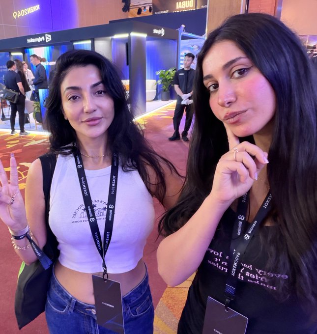 Two women posing at what appears to be a B8 Conference or event, both wearing lanyards, with one making a peace sign and the other resting her chin thoughtfully.