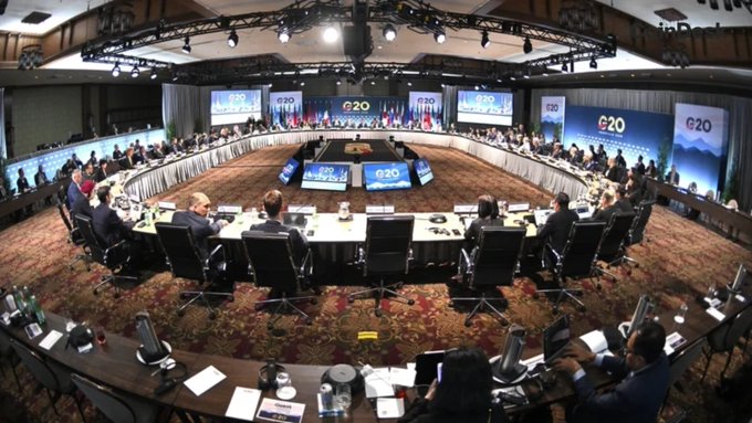 Delegates from G20 member nations gather around a large circular table at the G20 summit, with multiple screens displaying the "G20" logo, discussing key global priorities including digital assets, debt, and investment.