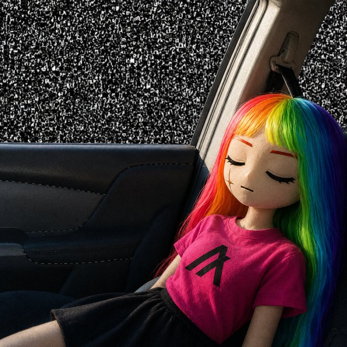 A handmade doll with long rainbow-colored hair sits inside a car, wearing a pink shirt with a black letter 'A' printed on it.