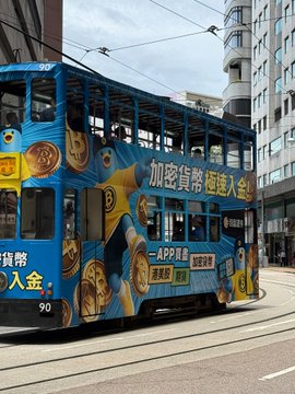 A blue double-decker tram in Hong Kong covered in cryptocurrency advertisements featuring cartoon coins and Chinese text promoting "加密貨幣" (cryptocurrency) and an APP download.