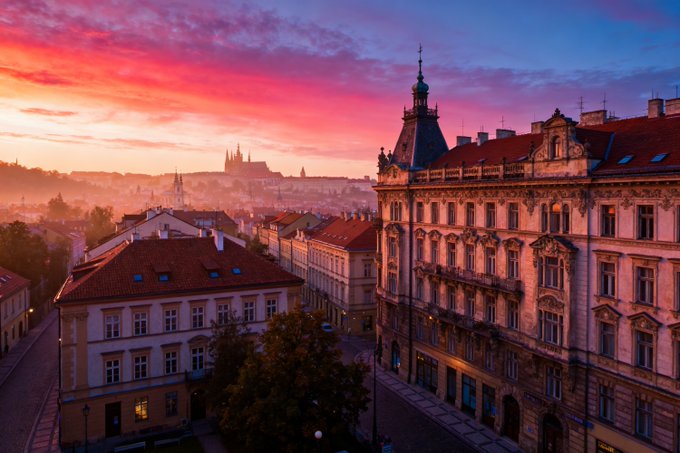 A scenic cityscape at sunset with a pink and orange sky, showing ornate architecture in the foreground and what appears to be Prague Castle in the misty distance.