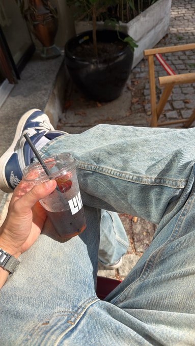 A first-person view shows someone holding an iced drink in a clear plastic cup with a black straw while sitting with crossed legs in jeans and blue and white sneakers.