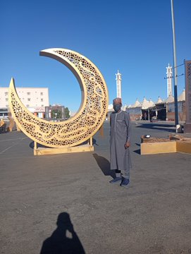 A man in traditional gray clothing stands beside a large golden crescent moon sculpture in an outdoor plaza with minarets visible in the background.