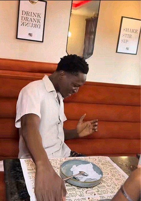 A man sits at a restaurant booth with an empty plate in front of him, gesturing with his hand while looking down, seemingly mid-conversation. The wall signs read "DRINK DRANK DRUNK" and "SHAKE STIR SIP," fitting the post's caption about being "positioned for ANSEM" with $2—suggesting a humorous context of having spent most of his money on a date or meal.