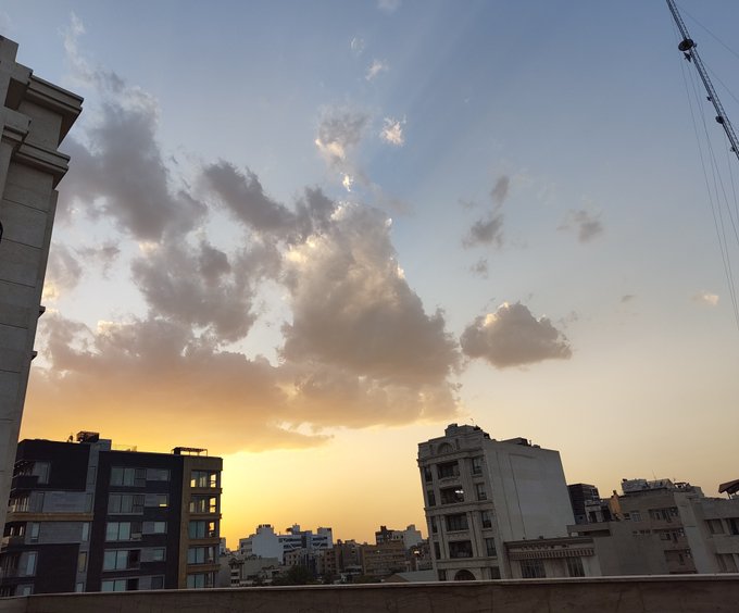 A dramatic sunset sky with golden rays piercing through scattered clouds above a city skyline frames the view, creating a serene backdrop for the long day's wait. The urban rooftops and construction crane in the foreground echo the patience of watching time pass, perfectly matching the "Day 5" countdown sentiment.