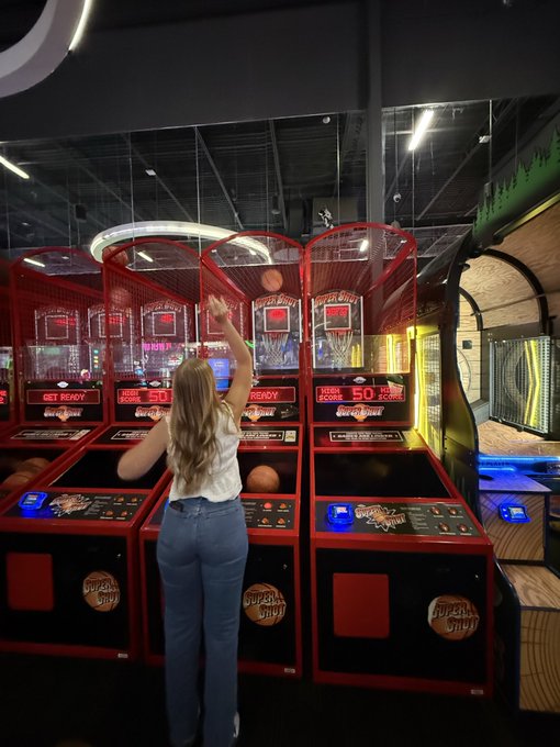 A woman with long blonde hair plays arcade basketball at a Pop-A-Shot machine in an entertainment venue, raising her arm to shoot.