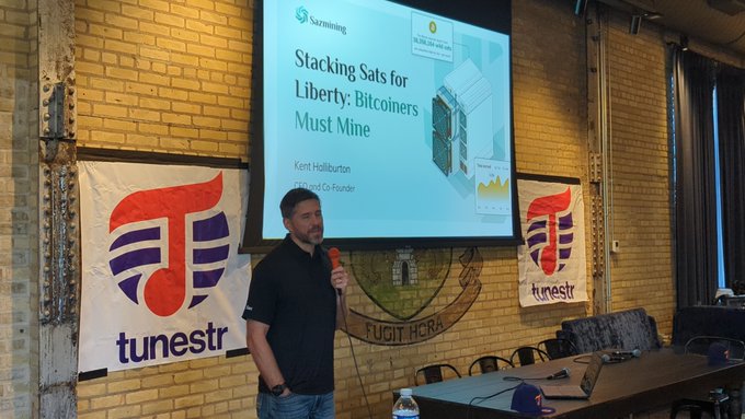A man stands on stage presenting 'Stacking Sats for Liberty: Bitcoiners Must Mine' at a Saxmining event, with 'tunestr' banners visible on either side of him.