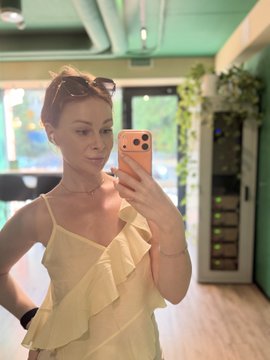 A woman in a yellow ruffled dress taking a mirror selfie with a gold iPhone in a teal-walled cafe or studio space.