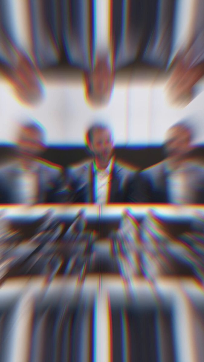 The image shows a group of people sitting around a table, possibly engaged in a meeting or discussion. The focus of the image is on one person who appears to be speaking at the front of the room. The background features a blurred image of other individuals seated behind them, suggesting that they are all part of the same gathering.