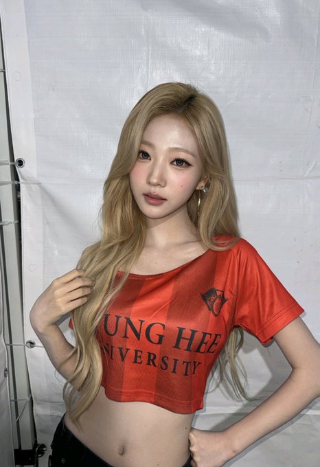 A young woman with long blonde hair poses wearing a red crop top that reads 'JUNG HEE UNIVERSITY' with a tiger logo on the chest.