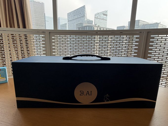 A black gift box with a handle, branded 'B.AI' with a moon and running figure design, placed on a wooden table near a window overlooking city buildings.