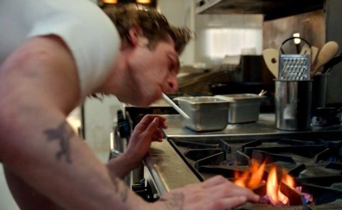 A young man with tattoos, wearing a white t-shirt, lights a cigarette in a commercial kitchen with his face close to flames bursting from a gas stove burner.