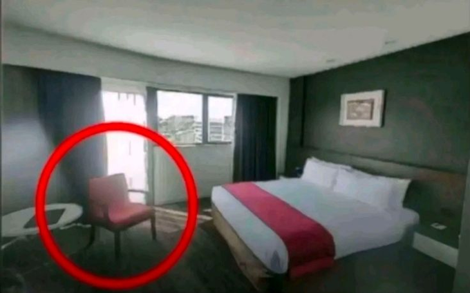 A hotel room with a bed featuring a red runner, where a red chair near the window has been circled in red.