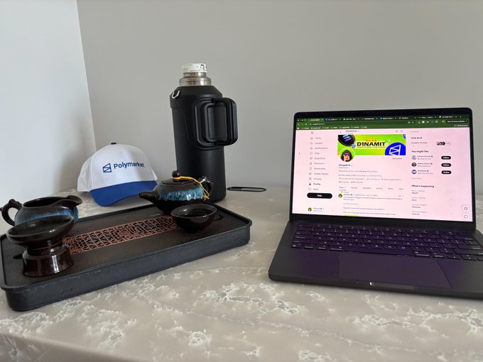 A tea set with a black and white "Polymarket" cap, a flask, and a laptop displaying a Polymarket page about "DINAMIT" sit on a table, perfectly capturing the "Tea session + X + Polymarket" vibes for a relaxed Monday.