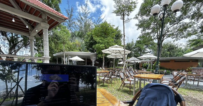 An outdoor cafe setting with wooden tables, chairs, and white umbrellas under trees, with a laptop and blue backpack visible in the foreground.