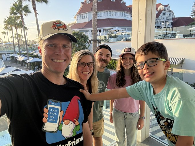 A group selfie of five people — two adults and three younger individuals — smiling outdoors at what appears to be a resort, with the man in front wearing a 'Phineas and Ferb' t-shirt reading 'If you know, you know.'