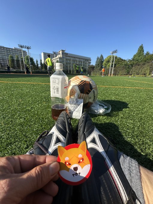 A first-person view at a soccer field showing black cleats, a soccer ball, a water bottle, and a hand holding a circular Shiba Inu (Doge) sticker.
