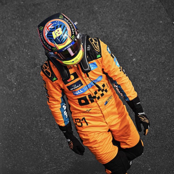 A racing driver wearing an orange racing suit with the number 81, 'DP World', 'Dell', and 'OKX' sponsor logos walks with head down, wearing a colorful Monster Energy branded helmet.