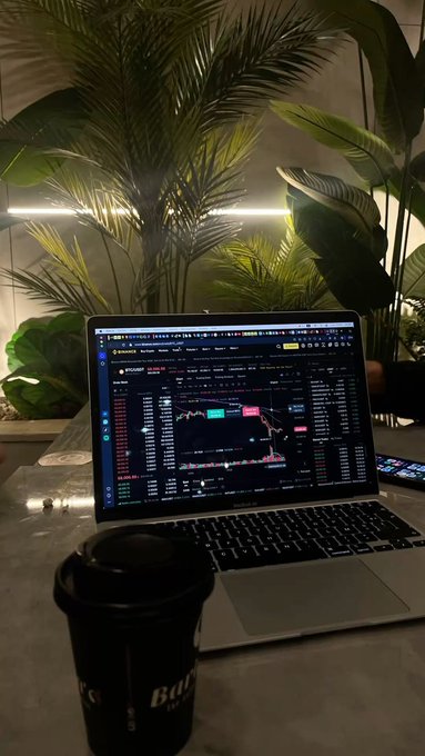 A laptop displaying a cryptocurrency trading chart sits on a table next to a black coffee cup, surrounded by tropical plants.