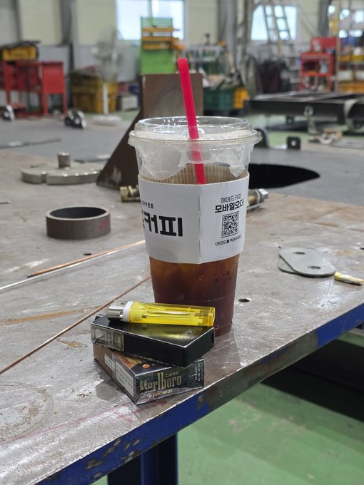 An iced coffee with a pink straw sits on a metal workbench alongside a yellow lighter and two stacked packs of Marlboro cigarettes, with Korean text and a QR code visible on the cup's sleeve—capturing the classic "altcoin investor's lunch" of caffeine and nicotine taken during a break in an industrial workshop setting.