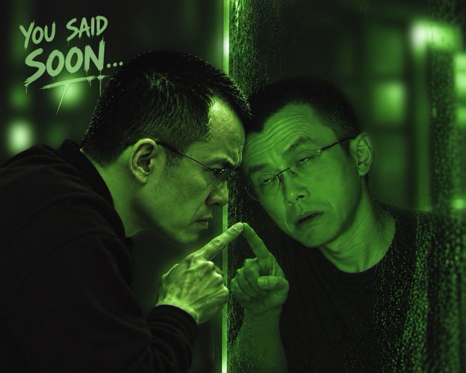 A man wearing glasses presses his forehead against a glass surface, staring at his reflection with a pointed finger. The text "YOU SAID SOON..." appears in green in the upper left corner.