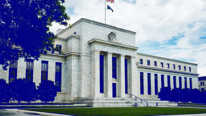 A photograph of the Federal Reserve building in Washington D.C. (Marriner Eccles Building) featuring its neoclassical architecture with prominent columns and an American flag on top, with the foreground tinted blue.