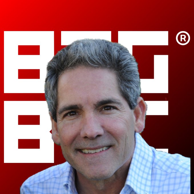 A headshot of a smiling, gray-haired man in a blue checkered shirt, set against a red background featuring a large white logo with a registered trademark symbol.