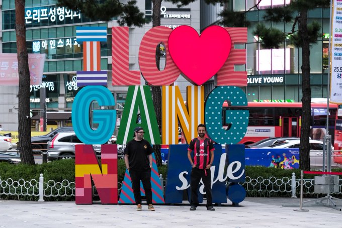 Two men stand in front of a large colorful 'I [heart] GANGNAM SEOUL' sign with Korean storefronts in the background