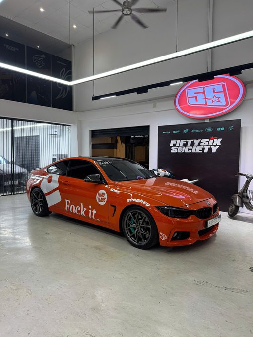 An orange BMW coupe with 'Fock it.' and 'Hard Heus' decals parked inside a 'FIFTYSIX SOCIETY' showroom.